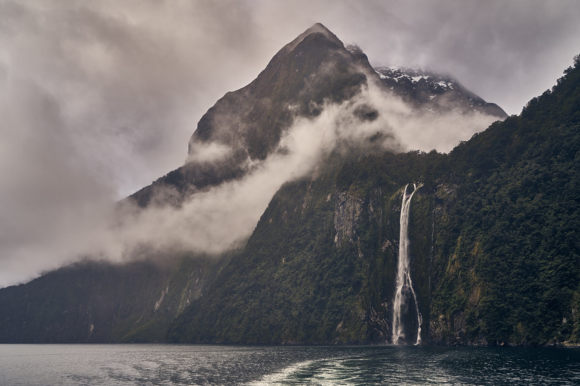 A cloudy day at Milford Sound, with a thin waterfall and mountain peaking through the clouds